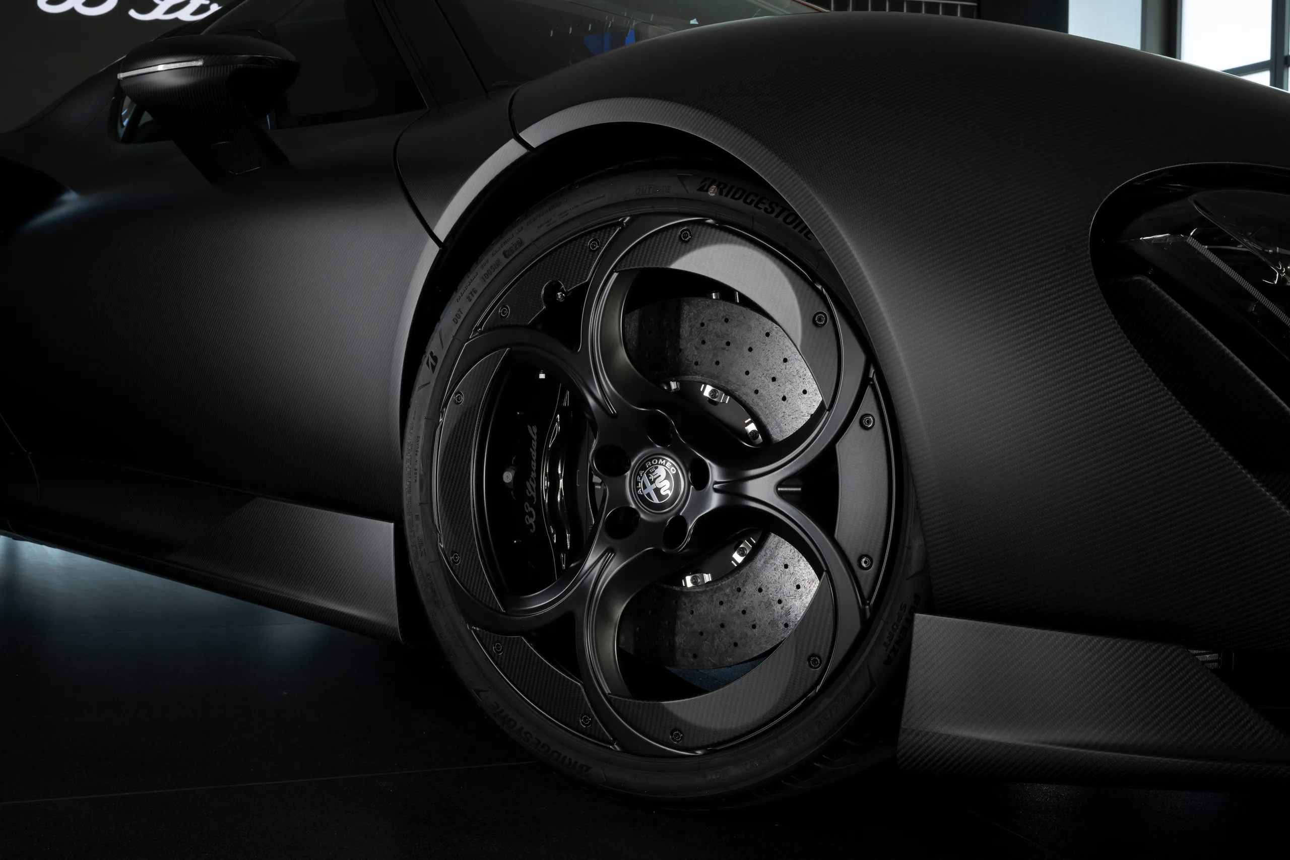 Close-up of a sleek black car wheel and tire on a dark background, showcasing intricate design and carbon fiber texture.